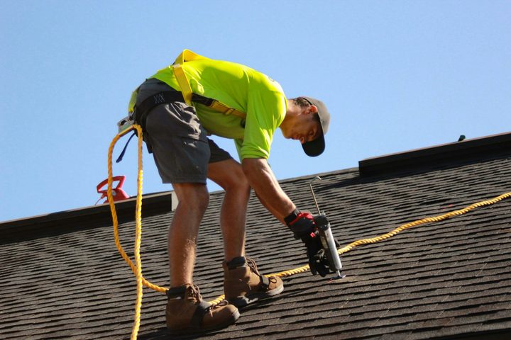 A roofer in a safety harness working on a shingle roof under a clear blue sky
